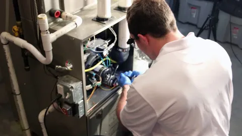 A technician evaluating furnace repair vs replacement works on the internal components of a gray metal furnace in a white shirt and blue gloves, using tools to repair the heating system.